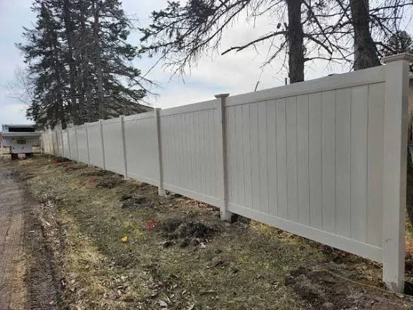 White vinyl privacy fence with vertical panels and concrete posts along tree line